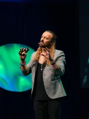 Anders Indset trägt ein schwarzes T-Shit, eine schwarze Hose und ein grau-meliertes Sakko. Er hat langes dunkelblondes, dass er sich halb hoch aus dem Gesicht gebunden hat. Er steht vor einem schwarzen Hintergrund mit blau-grünen dynamischen Blasen. Er gestikuliert.