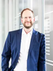 Carsten Wagner trägt einen mittelblauen Anzug mit weißem Hemd und braunen Gürtel. Er lehnt an einer Glaswand in einem modernem Büro. Er lächelt in die Kamera. Er hellbraunes kurzes Haar und er trägt eine Brille.