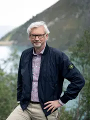 Michael Strugl trägt ein kariertes Hemd und eine leichte Outdoorjacke. Er lächelt in die Kamera. Im Hintergrund sehen wie Wiesen, Wälder und einen See. Er hat weißes welliges Haar und trägt eine Brille.