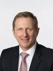 Alfons Haber trägt einen schwarzes Anzug mit einer rot-blau gemusterten Krawatte und ein weißes Hemd. Er hat dunkelblondes Haar und blaue Augen. Er lächelt.